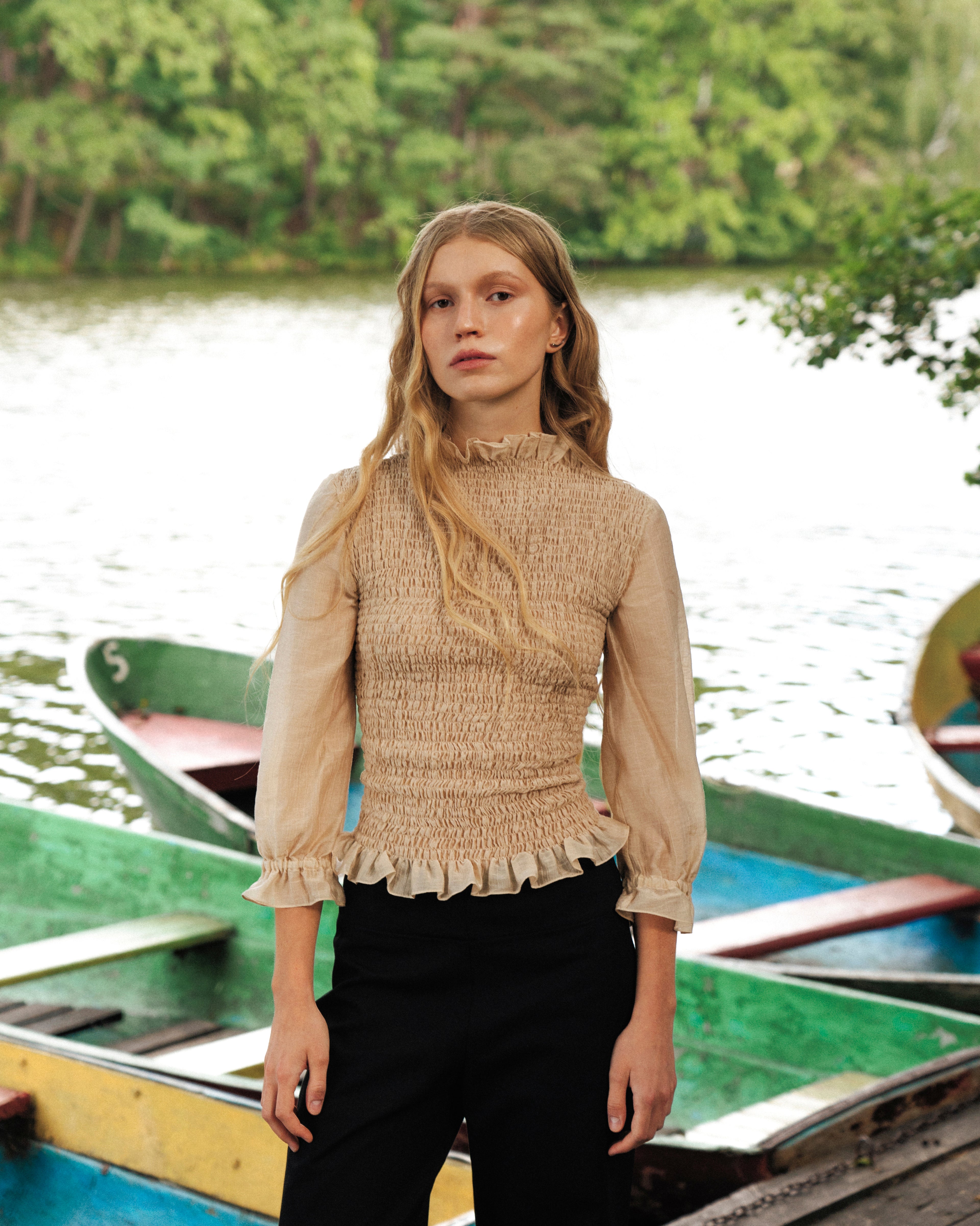 Woman in a beige blouse 3/4 sleeves standing by a lake with boats in the background