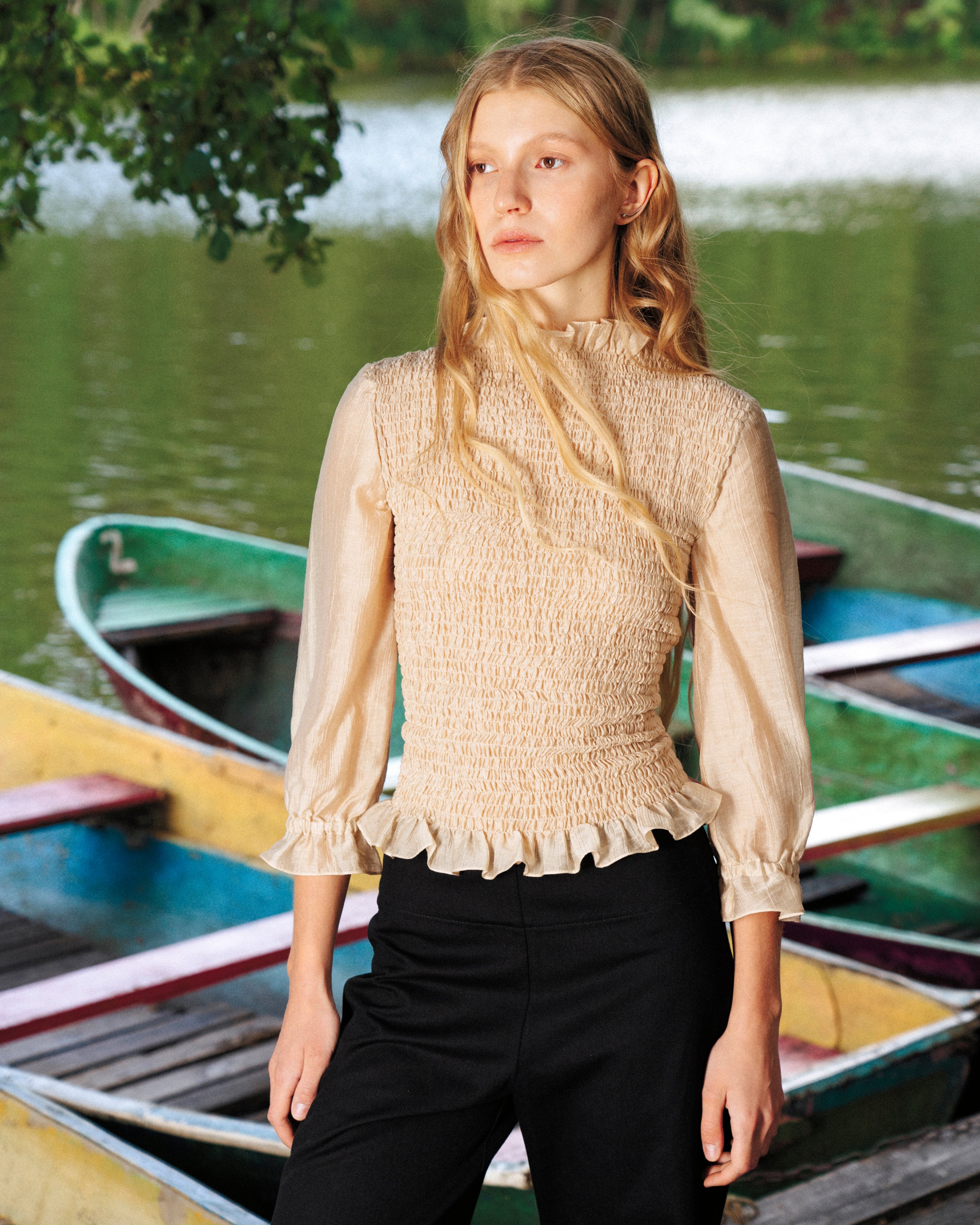 Woman in a beige blouse 3/4 sleeves and black pants standing near rowboats on a lake.
