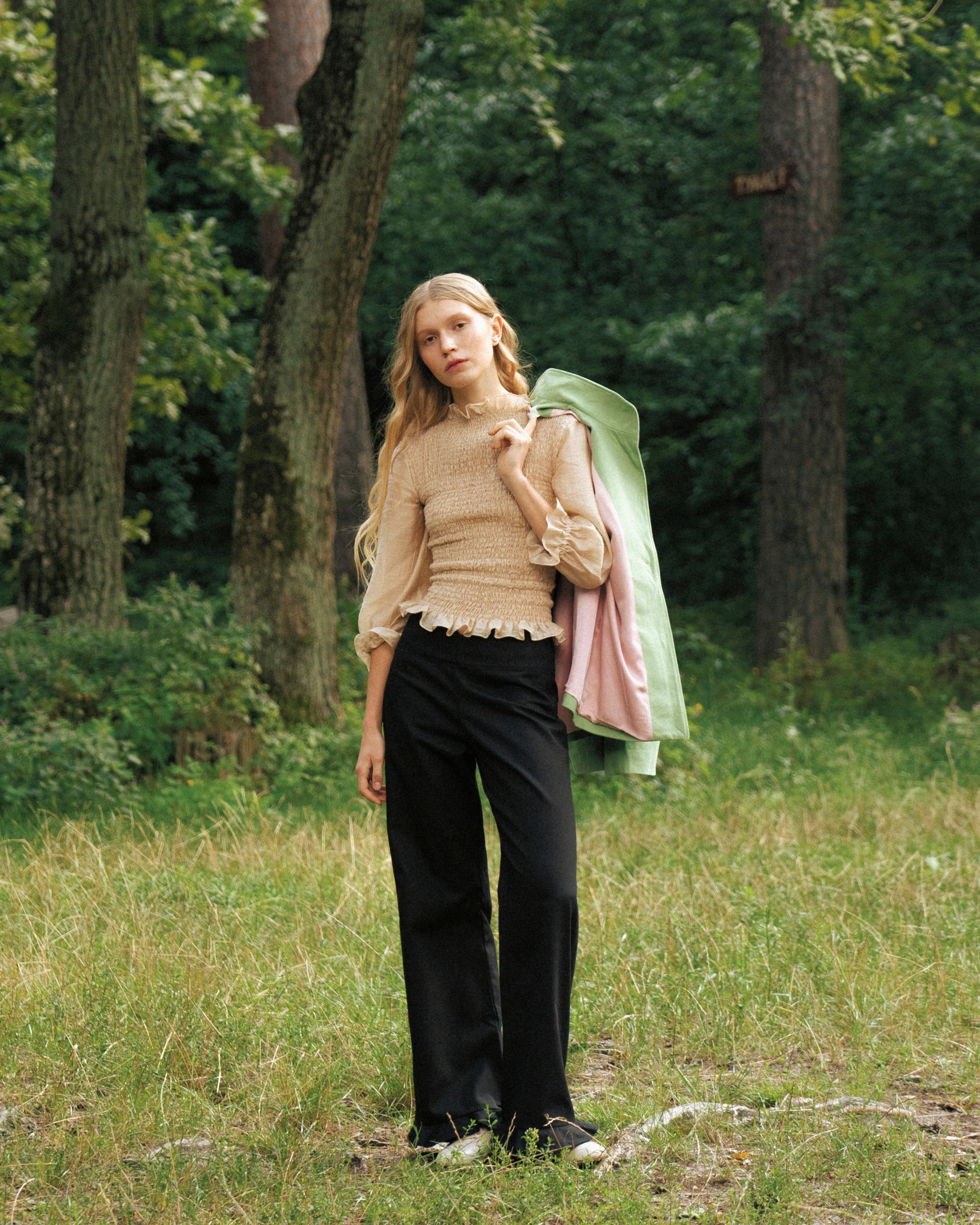 Woman standing in a forest with a green and pink jacket draped over her shoulder, beige blouse 3/4 sleeves and black cotton pants
