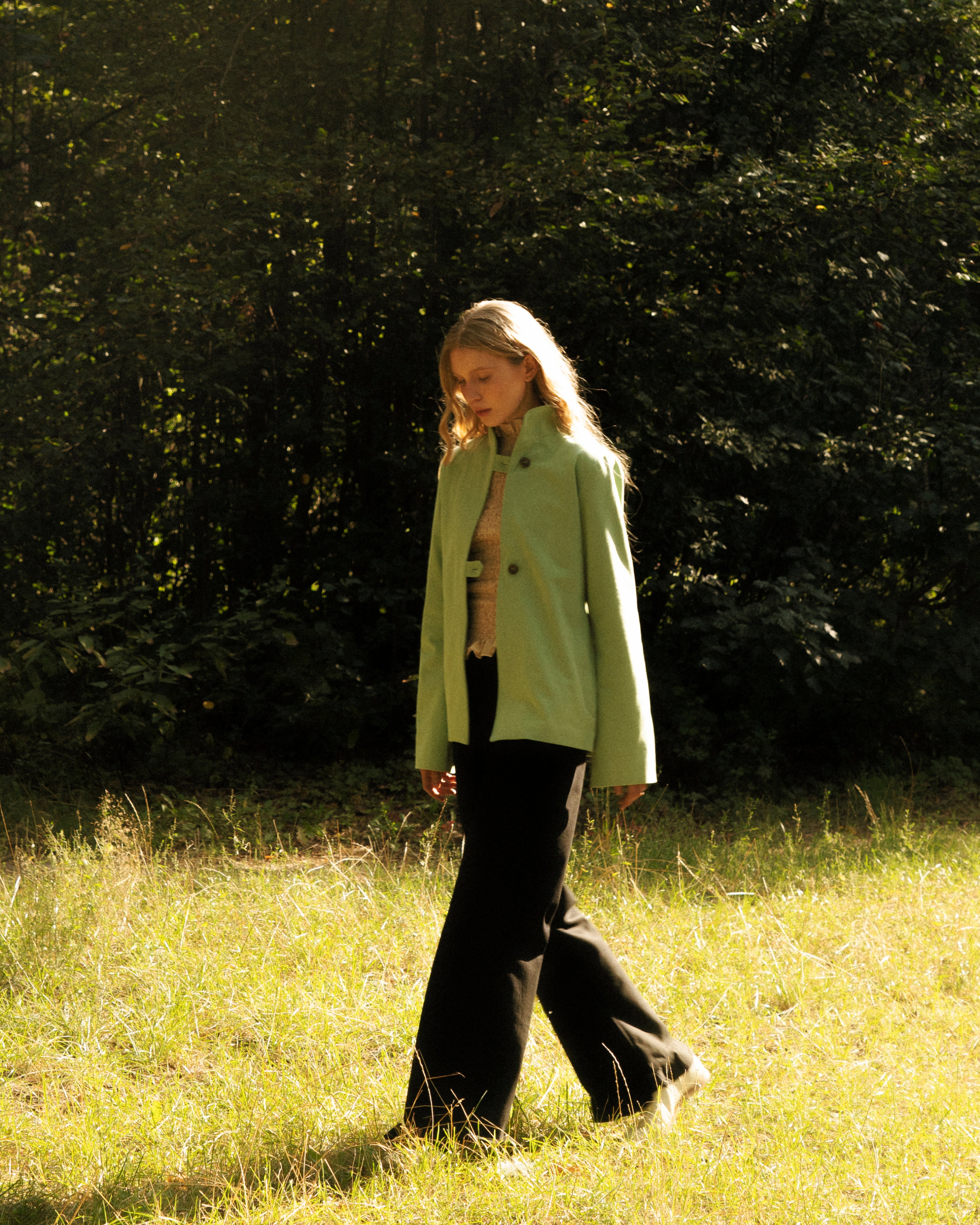 Woman in a light green jacket walking through a grassy field with trees in the background