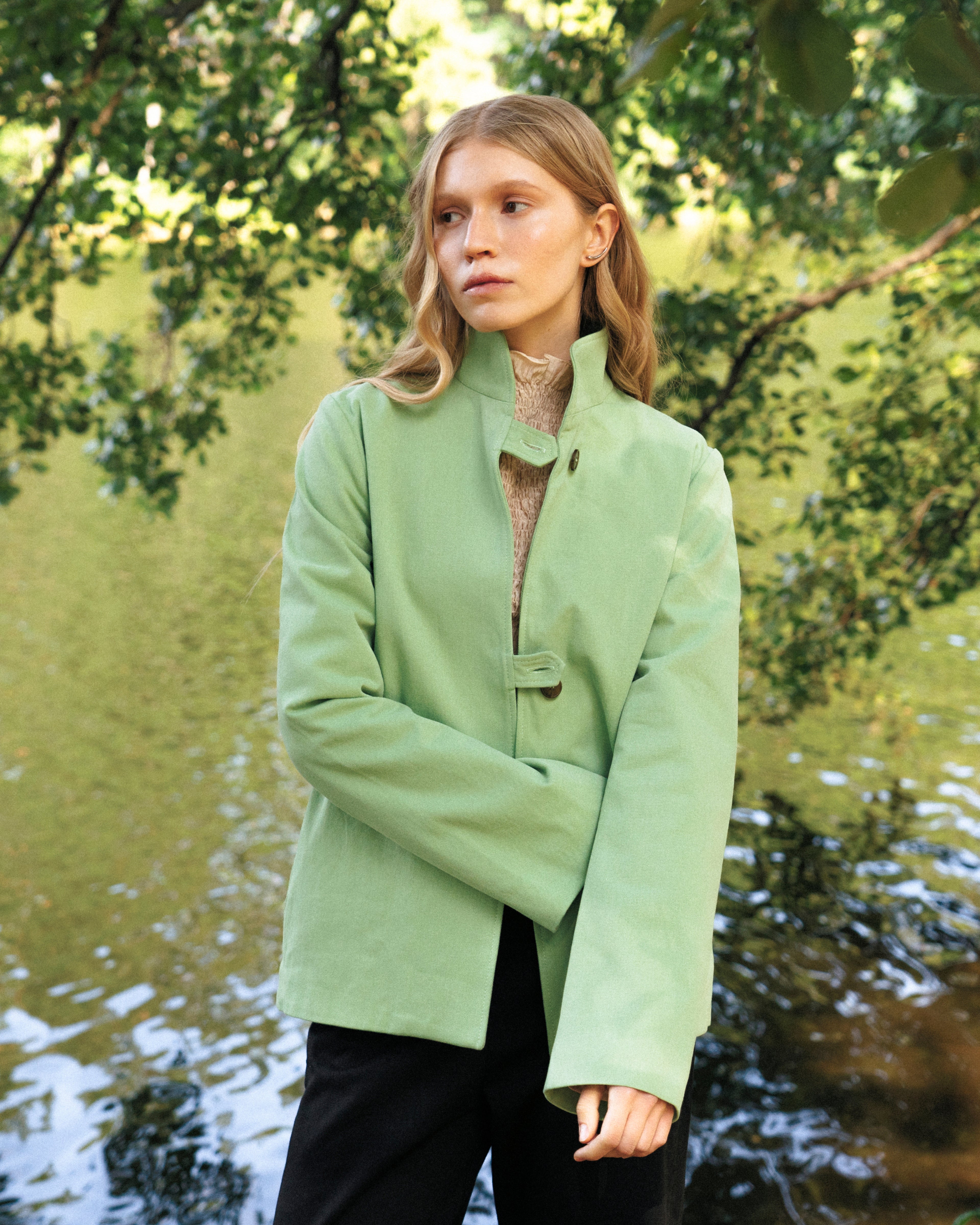 Woman wearing a light green jacket standing by water with trees in the background