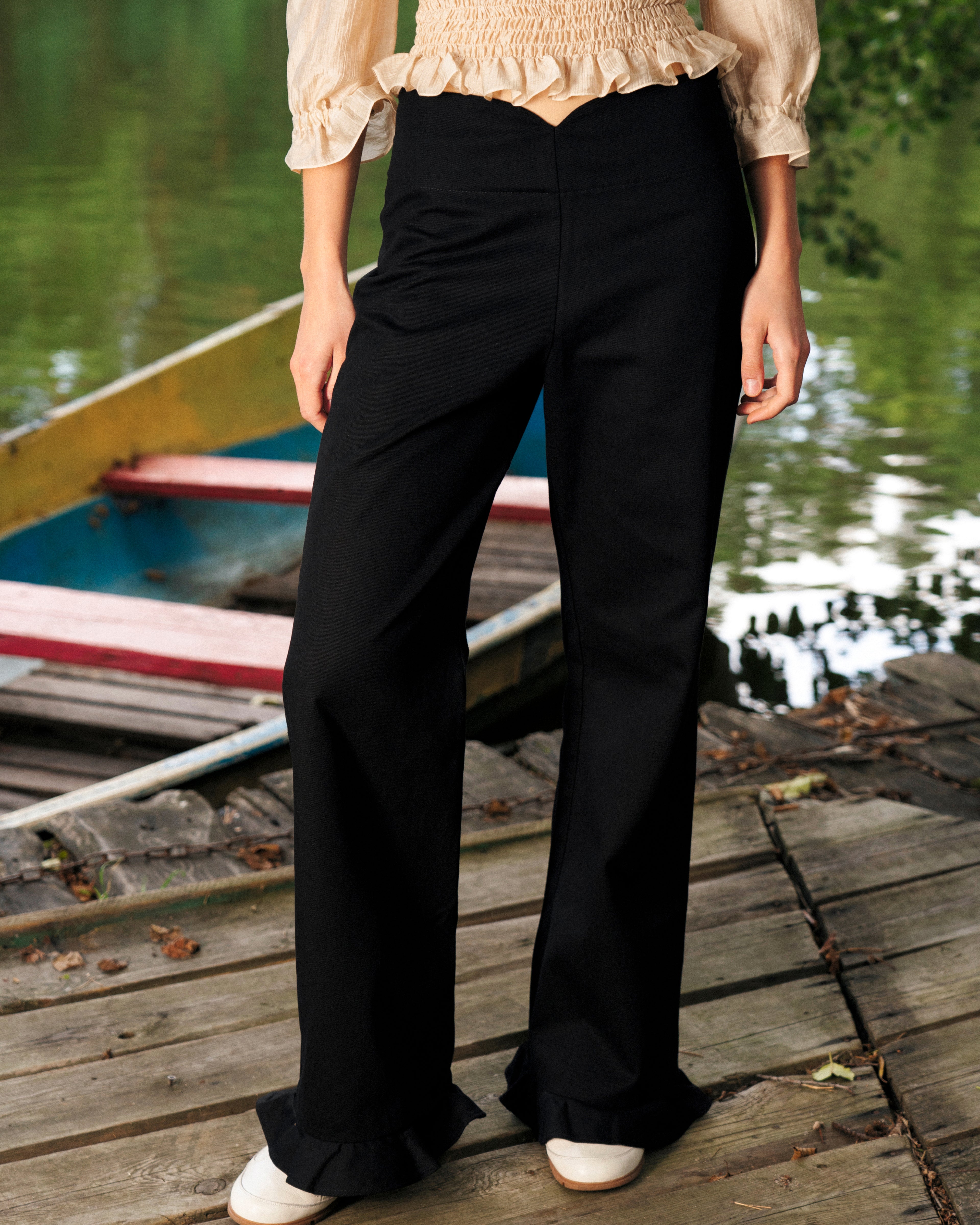 Person wearing black pants with ruffles and a beige top on a wooden dock with boats in the background.