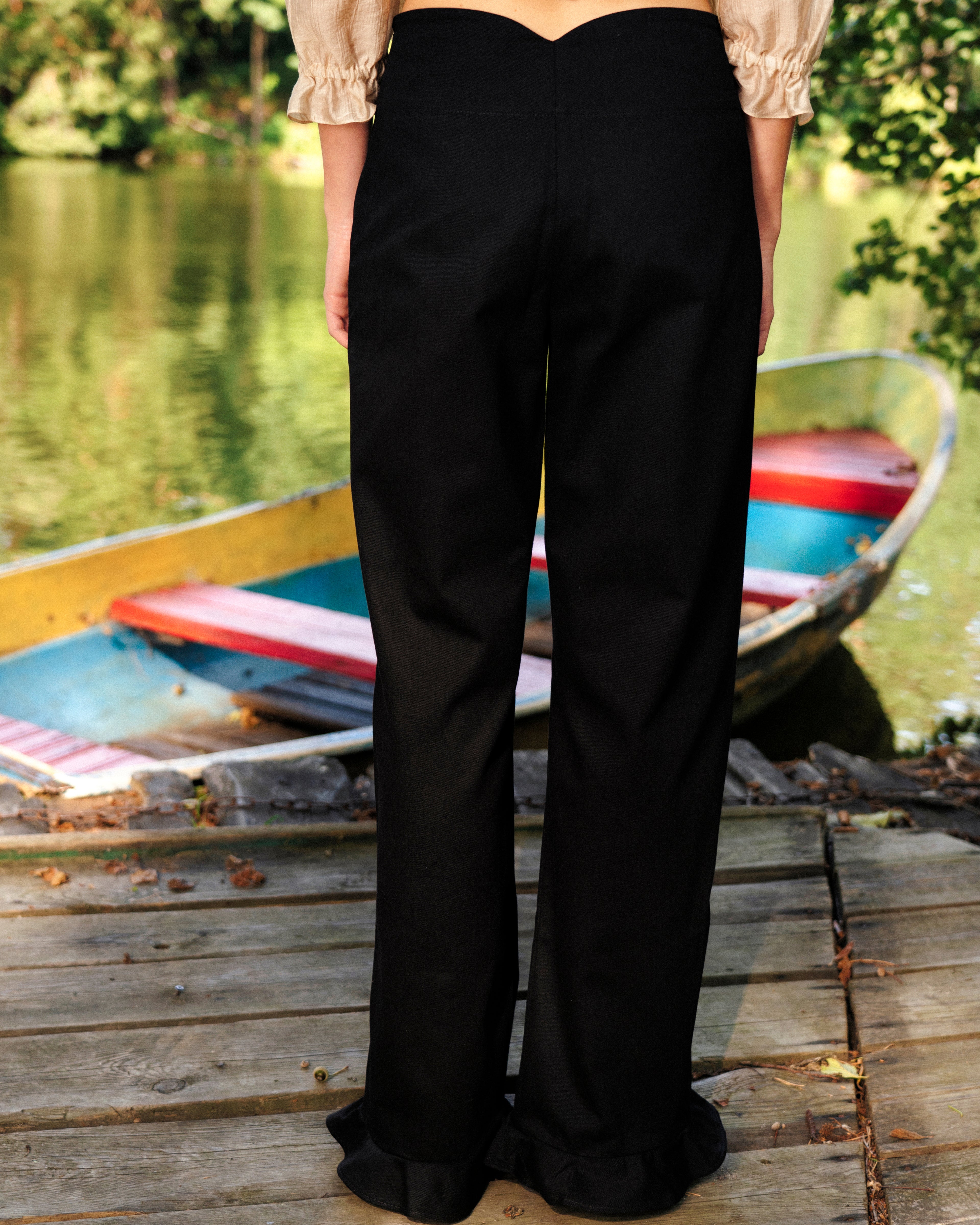 Person wearing black cotton pants with ruffles standing on a wooden dock with boats in the background