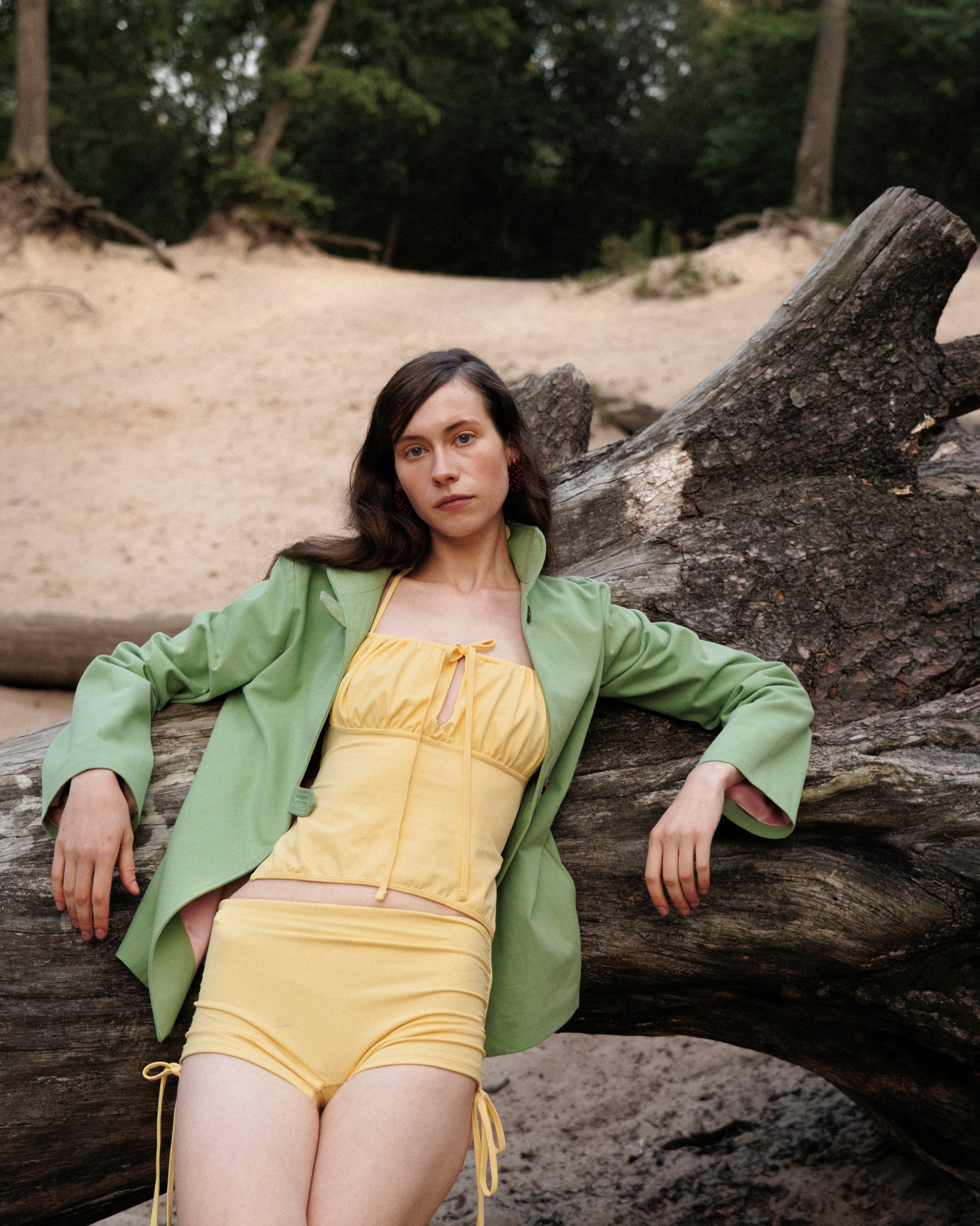 Woman in yellow tankini, swimwear shorts and green jacket sitting on a log with a natural background