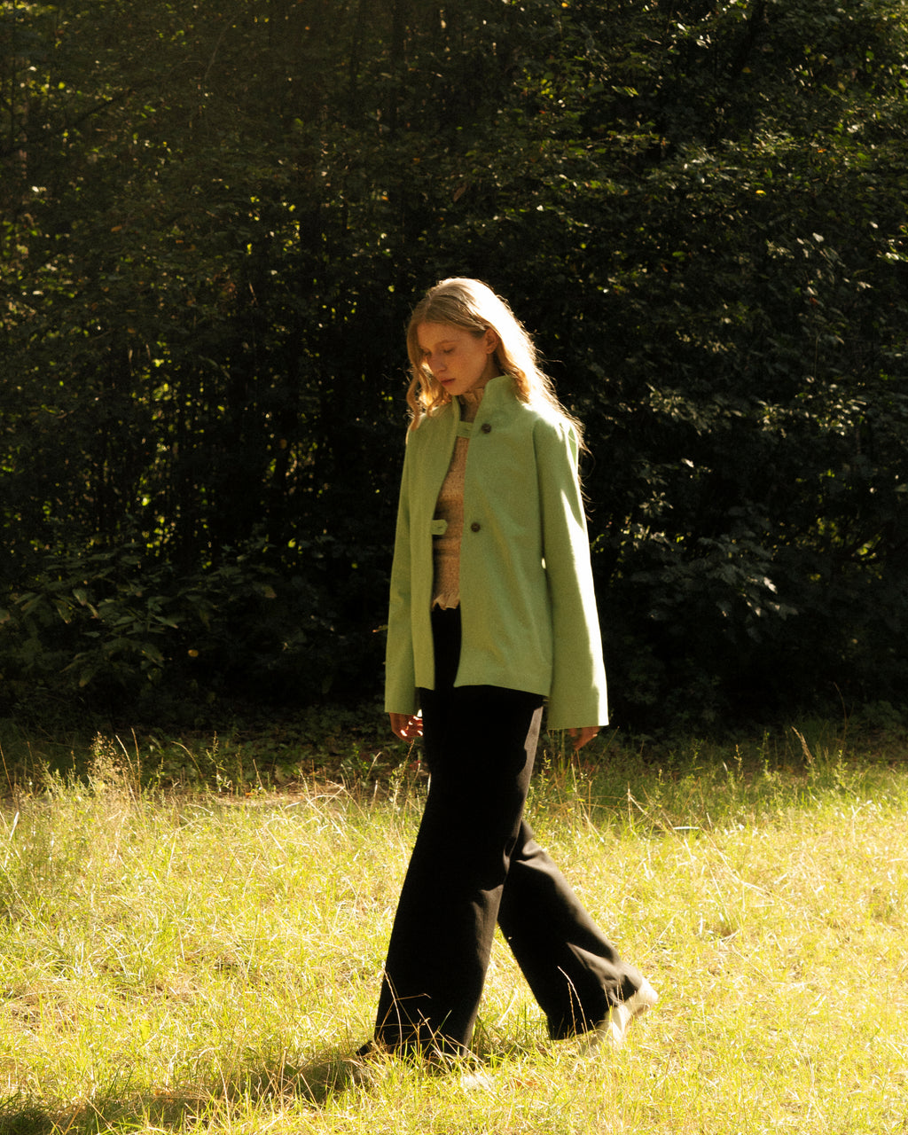 Woman in a light green jacket walking through a grassy field with trees in the background